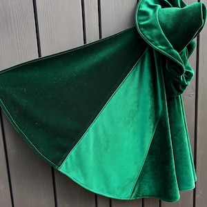 May include: A dark green velvet cloak with a hood. The cloak has a triangular design with two shades of green. The hood is gathered and has a rounded shape. The cloak is made of a soft, plush material.