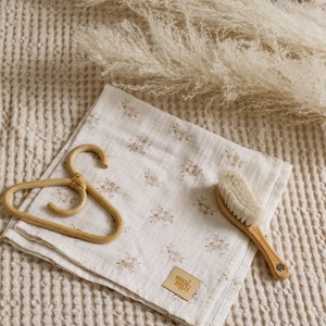 May include: A white muslin baby blanket with a floral print and a brown leather tag that says "moi nuli". The blanket is folded and lying on a beige waffle weave fabric with a wooden hanger and a wooden baby brush.