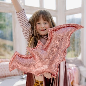 Op de afbeelding: Een roségouden, vleermuisvormig pluche kussen met lovertjes, vastgehouden door een kind in een gestreept shirt met lange mouwen en een mauve fluwelen cape. Het vleermuiskussen heeft een glinsterend, getextureerd oppervlak met lovertjes.