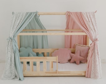 Puede incluir: Una cama de madera para niños pequeños con dosel de tela azul claro y rosa. La cama tiene una barandilla de seguridad y está decorada con almohadas.