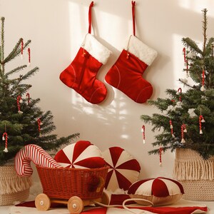 May include: A red and white candy cane themed Christmas scene with two red stockings hanging on a white wall, two small Christmas trees decorated with candy canes, a red and white striped candy cane cushion, a red and white striped candy cane wagon, and two red and white peppermint swirl cushions.