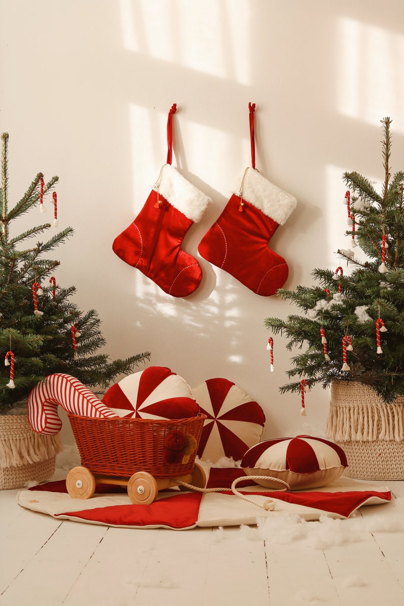 May include: A red and white candy cane themed Christmas scene with two red stockings hanging on a white wall, two small Christmas trees decorated with candy canes, a red and white striped candy cane cushion, a red and white striped candy cane wagon, and two red and white peppermint swirl cushions.