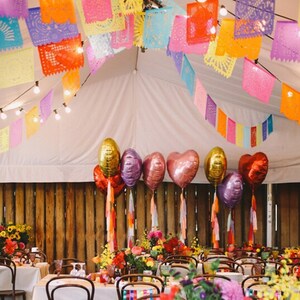 May include: A wedding reception venue decorated with colorful paper lanterns and balloons. The tables are set with white tablecloths and floral centerpieces. The chairs are decorated with colorful ribbons. The venue is decorated with a rustic wooden wall and a white tent.