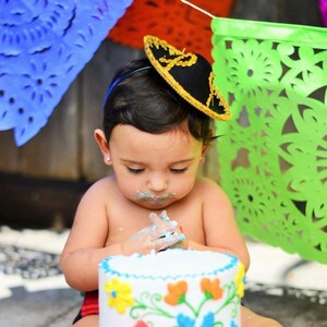 May include: A baby wearing a sombrero headband is sitting in front of a white cake decorated with colorful flowers and a red heart. The cake is on a glass cake stand.