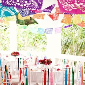 May include: White chairs with colorful ribbons tied to the back, set for a wedding reception. The chairs are arranged around a white tablecloth with flowers on the table. Colorful paper decorations hang from the ceiling.