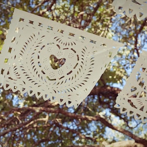 May include: White paper cutouts with intricate designs hang from a tree branch. The cutouts are shaped like squares with rounded corners and feature a heart shape in the center.