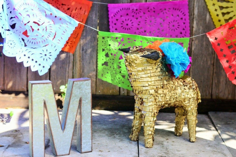 May include: A gold pinata shaped like a bull with blue and orange paper decorations. The pinata is sitting on a wooden surface in front of a wooden wall with a string of colorful paper flags hanging above it. The letter "M" is in the foreground.