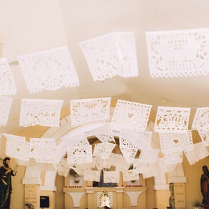 May include: White paper decorations with intricate cutouts hang from the ceiling of a church. The decorations are in the shape of squares and are strung together in a line. The decorations are a traditional Mexican paper craft.