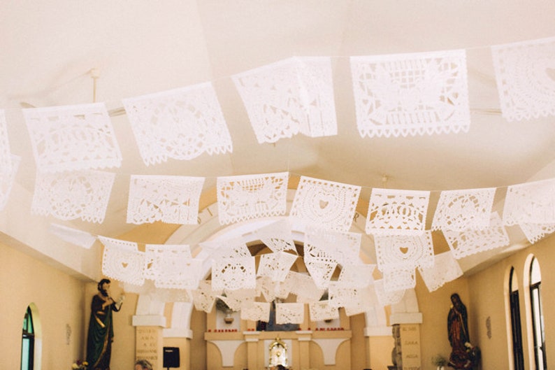 May include: White paper decorations with intricate cutouts hang from the ceiling of a church. The decorations are in the shape of squares and are strung together in a line. The decorations are a traditional Mexican paper craft.