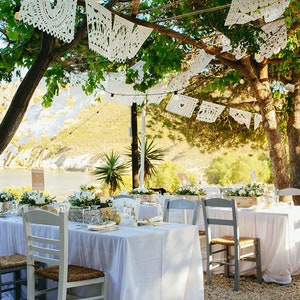 May include: A wedding reception table setting with white tablecloths and white chairs under a tree decorated with white paper lanterns. The table is set with white flowers and greenery.