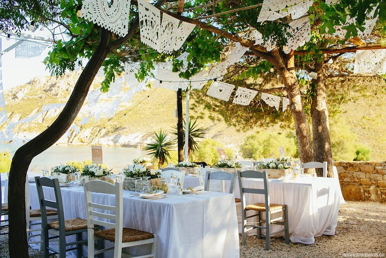 May include: A wedding reception table setting with white tablecloths and white chairs under a tree decorated with white paper lanterns. The table is set with white flowers and greenery.