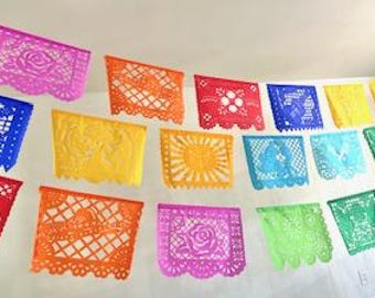 May include: A colorful string of papel picado banners in various colors, including pink, orange, red, blue, yellow, and green. Each banner features intricate cut-out designs, such as flowers, geometric patterns, and suns. The banners are hanging against a white wall.