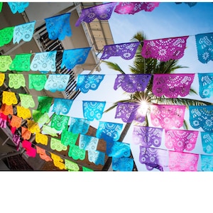 May include: A vibrant display of colorful papel picado banners strung across a street. The banners are in shades of pink, purple, blue, green, yellow, and orange, with intricate cut-out designs. The sun shines through a palm tree in the background.