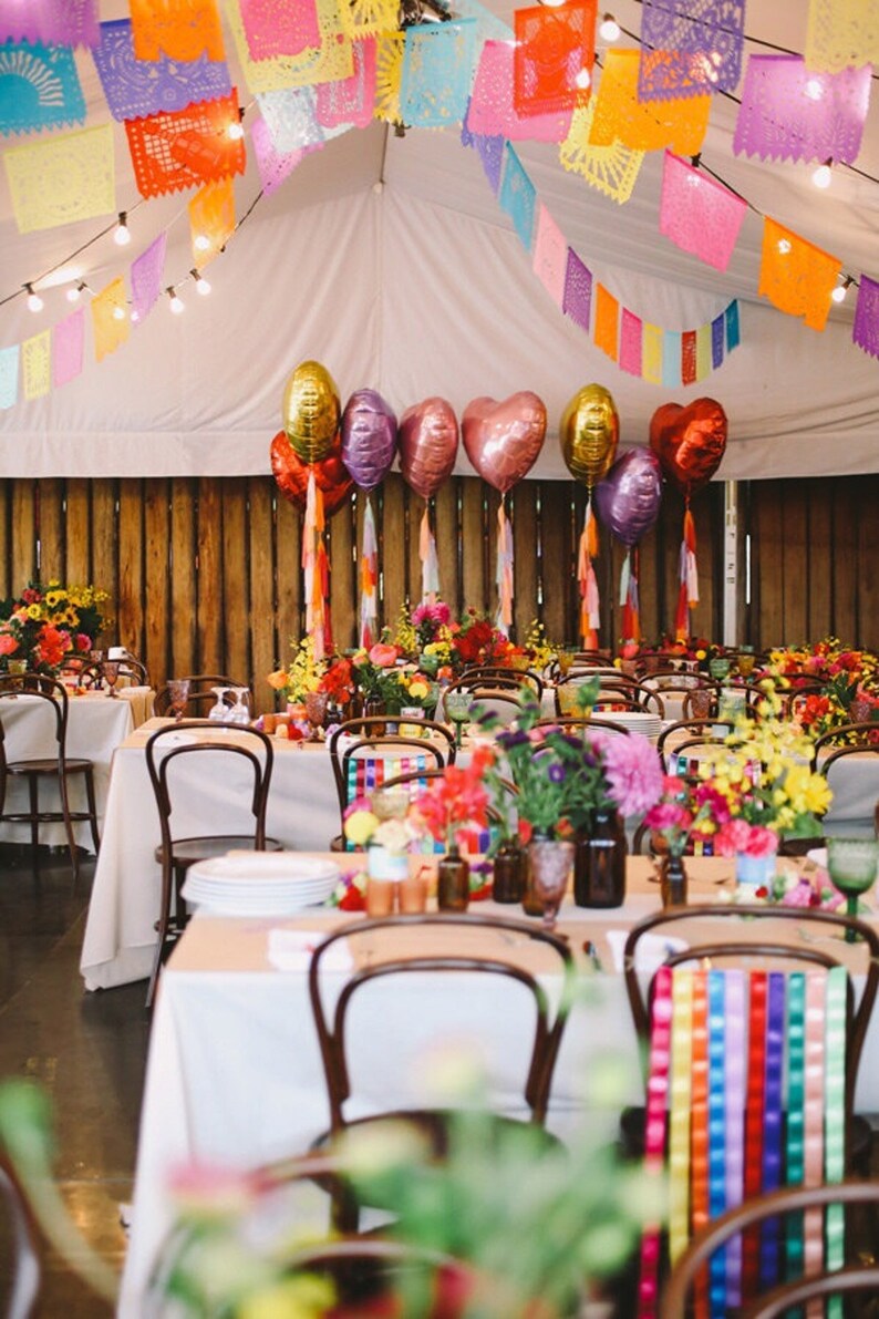 May include: A wedding reception venue decorated with colorful paper lanterns and balloons. The tables are set with white tablecloths and floral centerpieces. The chairs are decorated with colorful ribbons. The venue is decorated with a rustic wooden wall and a white tent.