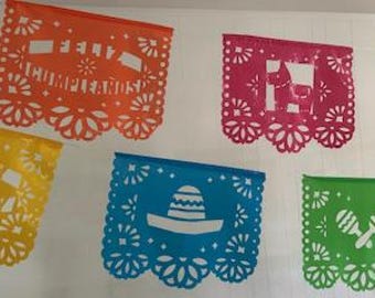 May include: Colorful papel picado banners in orange, pink, yellow, blue, and green hang against a white background. The banners feature cut-out designs of a sombrero, maracas, and the text "FELIZ CUMPLEAÑOS".