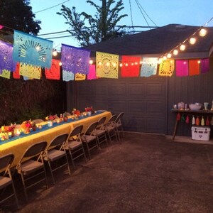 May include: A yellow tablecloth covers a long table with folding chairs set for a party. Colorful paper decorations hang from a string of lights above the table.