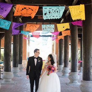May include: A wedding scene with a couple walking under colorful papel picado banners. The bride wears a white dress and veil, holding a bouquet. The groom is in a black tuxedo. The setting is a covered walkway with wooden pillars.