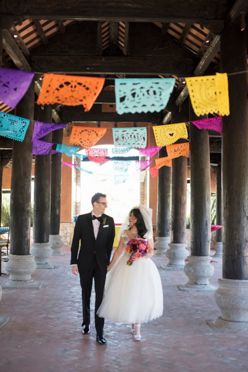 May include: A bride and groom walking under a colorful paper banner. The bride is wearing a white dress and a bouquet of flowers. The groom is wearing a black suit and a white bow tie.