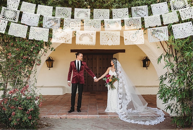 May include: A couple holds hands in front of a white building decorated with white paper cutouts. The groom is wearing a burgundy velvet suit jacket and a black bow tie. The bride is wearing a white lace wedding dress and a long white veil.