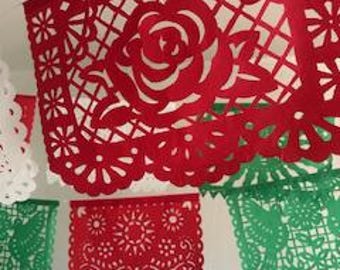 May include: Red, white, and green papel picado banners with intricate cut-out designs. The banners feature floral and geometric patterns, including a prominent rose. The festive decorations are strung against a white wall.