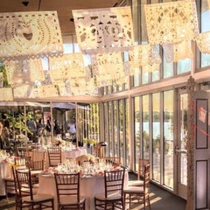 May include: White paper decorations, called "papel picado", hang from the ceiling of a room with large windows. The room is decorated with tables and chairs for a party or event.