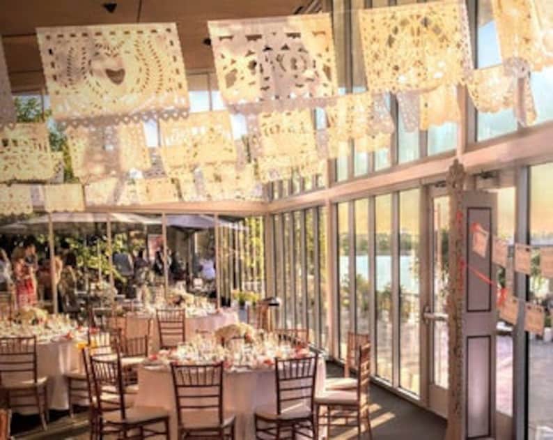 May include: White paper decorations, called "papel picado", hang from the ceiling of a room with large windows. The room is decorated with tables and chairs for a party or event.