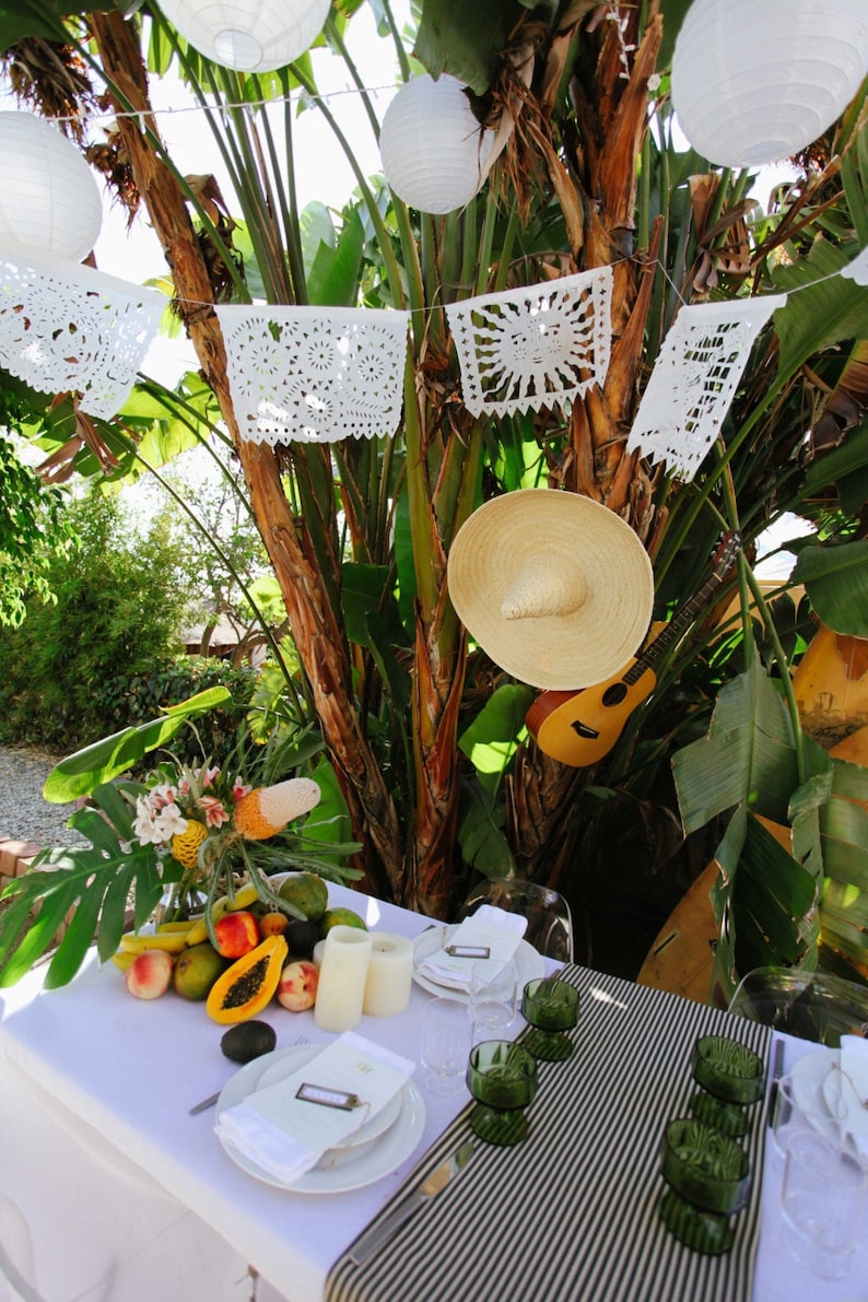 May include: A white tablecloth with a black and white striped runner covers a table set for a party. The table is decorated with a centerpiece of fruit, a straw hat, and a guitar. White paper lanterns hang from the branches of a palm tree overhead.