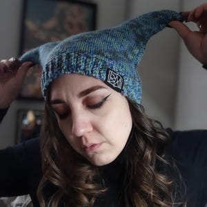 May include: A blue and green knit hat with a pointed top. The hat has a small black and white tag on the front. The hat is being held up by a person.