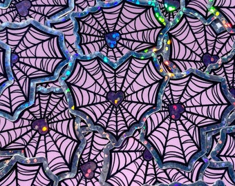 May include: A sheet of holographic stickers featuring a repeating pattern of black spiderwebs with hearts in the center. The stickers have a silver iridescent finish.