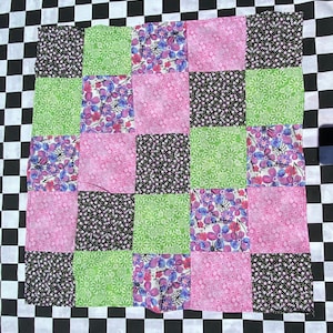 May include: A patchwork quilt with a checkerboard border. The quilt features a grid of squares in various floral patterns and colors, including pink, green, black, and purple. The quilt is handmade and has a cozy, vintage aesthetic.