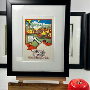 May include: Framed art print of a colorful landscape scene with the text "Van Briggle Art Pottery Colorado Springs Colo." The print is in a black frame with a white mat, displayed on a small wooden easel. A red apple sits nearby.