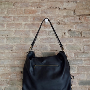 Op de afbeelding: Een zwarte leren tote bag met gouden hardware hangend aan een bakstenen muur.