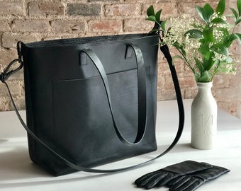May include: A black leather tote bag with a long strap and a front pocket. The bag is sitting on a white surface next to a pair of black leather gloves.