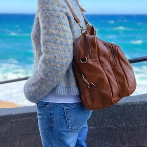 May include: A brown leather backpack with multiple compartments and gold-colored hardware. The backpack is worn over the shoulder. The person is wearing a blue and yellow sweater and blue jeans. The background is a view of the ocean.