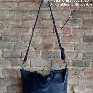 May include: A dark blue leather tote bag with two top handles and a removable crossbody strap. The strap is adjustable and has a gold metal clasp. The bag is hanging on a white hook against a brick wall.