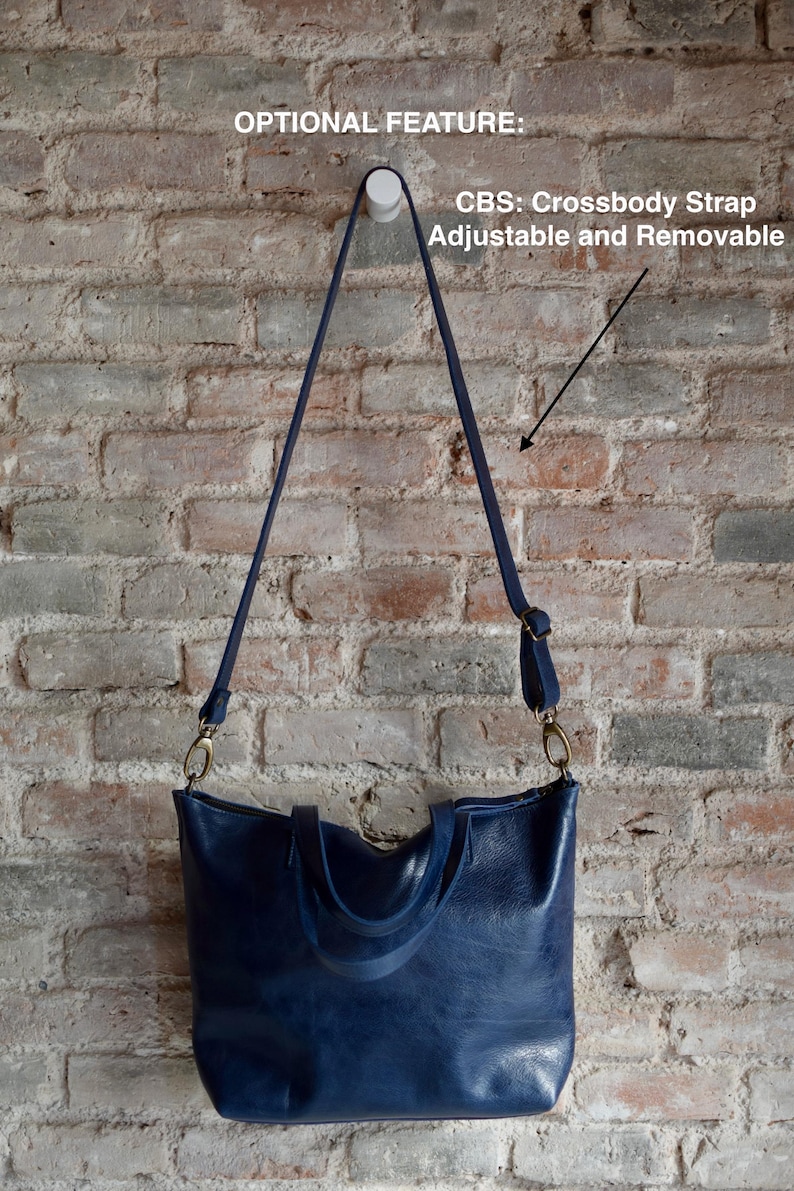 May include: A dark blue leather tote bag with two top handles and a removable crossbody strap. The strap is adjustable and has a gold metal clasp. The bag is hanging on a white hook against a brick wall.