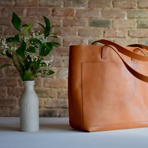 May include: A large, brown leather tote bag with a single pocket on the front. The bag has two long straps and is sitting on a white surface.