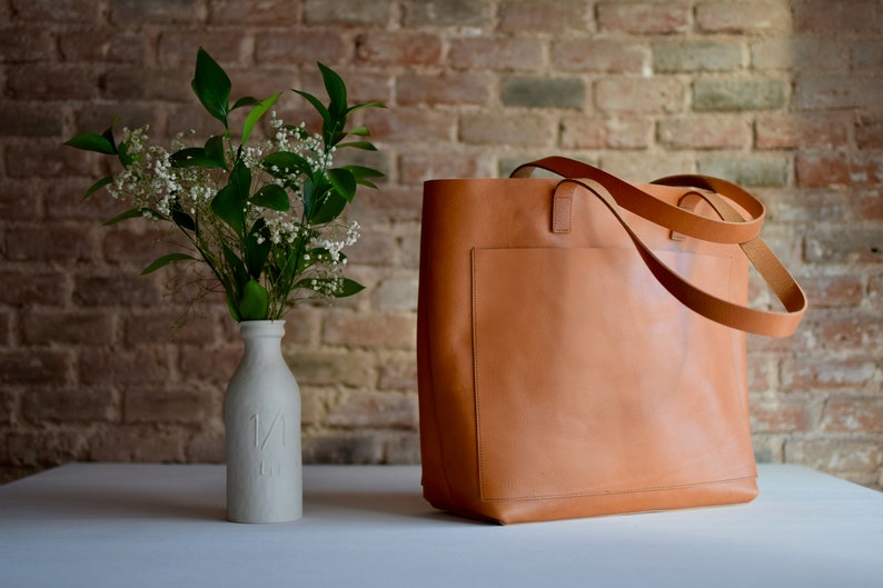 May include: A large, brown leather tote bag with a single pocket on the front. The bag has two long straps and is sitting on a white surface.