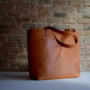 May include: A brown leather tote bag with a single pocket on the front. The bag has two long straps.