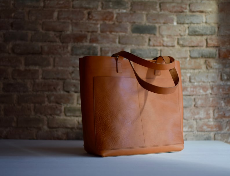May include: A brown leather tote bag with a single pocket on the front. The bag has two long straps.
