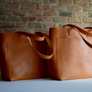 May include: Two brown leather tote bags with a front pocket. The bags are sitting on a white surface.