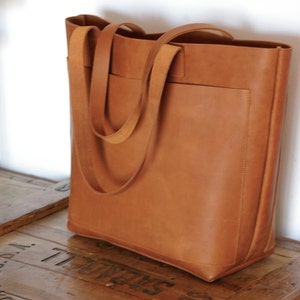 May include: A large, brown leather tote bag with two long straps. The bag has a large pocket on the front.