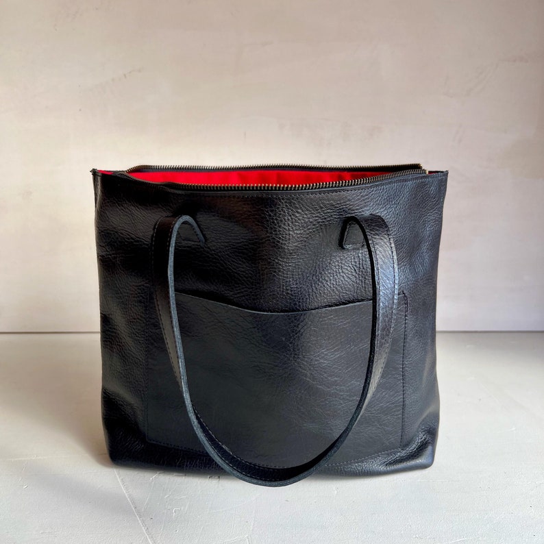 May include: Black leather tote bag with a red lining and a zipper closure. The bag has two top handles and a front pocket.