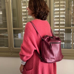 Peut inclure: Un sac à dos en cuir bordeaux profond avec une finition lisse, une poignée supérieure et une poche frontale zippée horizontale dotée d'une tirette en cuir assortie. Le sac est porté avec de fines bretelles réglables sur un pull en maille rose.