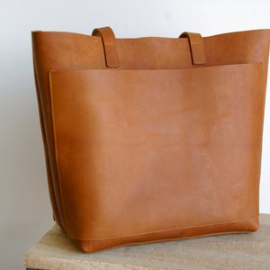 May include: A large, brown leather tote bag with a single, large pocket on the front. The bag has two leather straps.