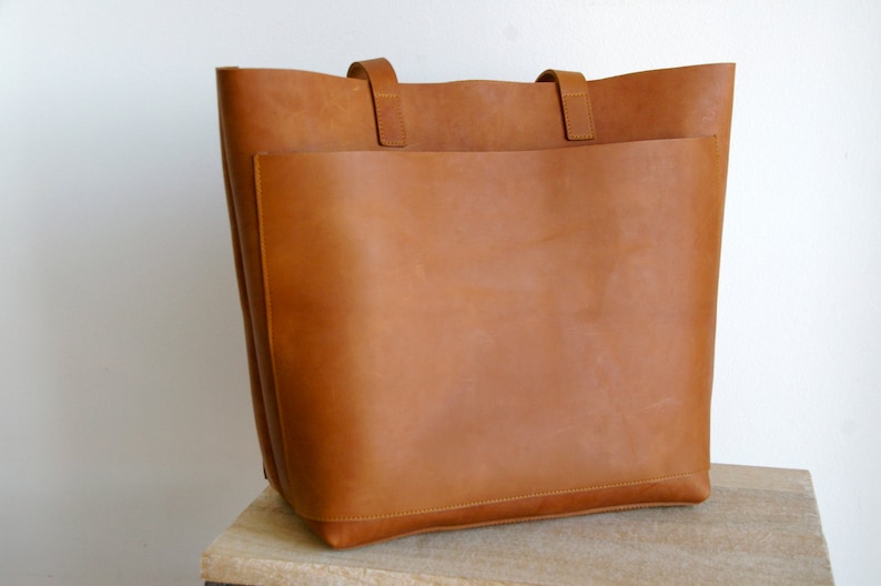 May include: A large, brown leather tote bag with a single, large pocket on the front. The bag has two leather straps.