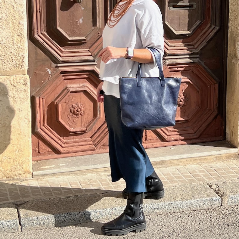 May include: A woman wearing a white sweater and a blue skirt is carrying a dark blue leather tote bag with a zipper closure. She is wearing black ankle boots.