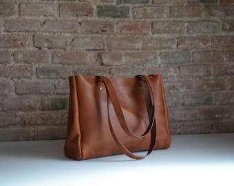 May include: A brown leather tote bag with two straps. The bag is sitting on a white surface in front of a brick wall.