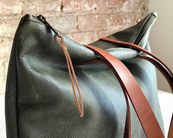 Può includere: Una borsa tote in pelle verde con cinturini in pelle marrone e chiusura a zip. La borsa ha un piccolo anello in pelle attaccato alla zip.