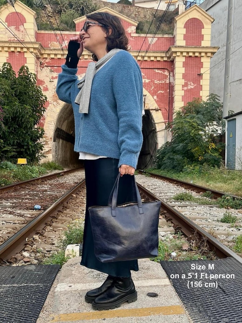 May include: A blue cardigan with a gray scarf worn by a person standing on a railroad track. The person is holding a black leather tote bag. Size M on a 5'1" person (156 cm).